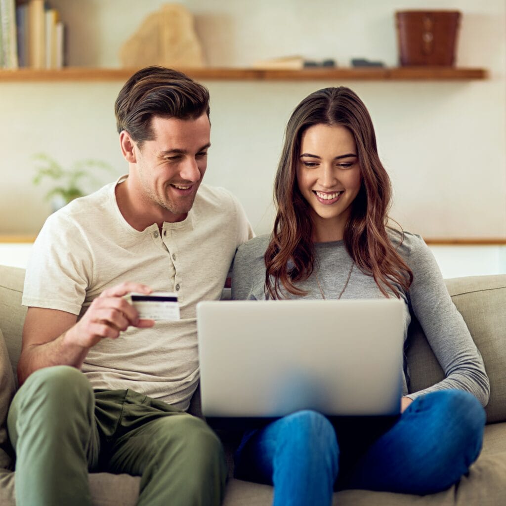 Man and woman sitting on a couch. The man is holding a credit card, and the woman has a computer on her lap.