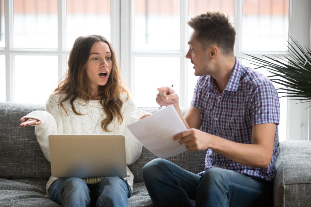 Couple sitting on a couch holding papers and having a tense discussion.